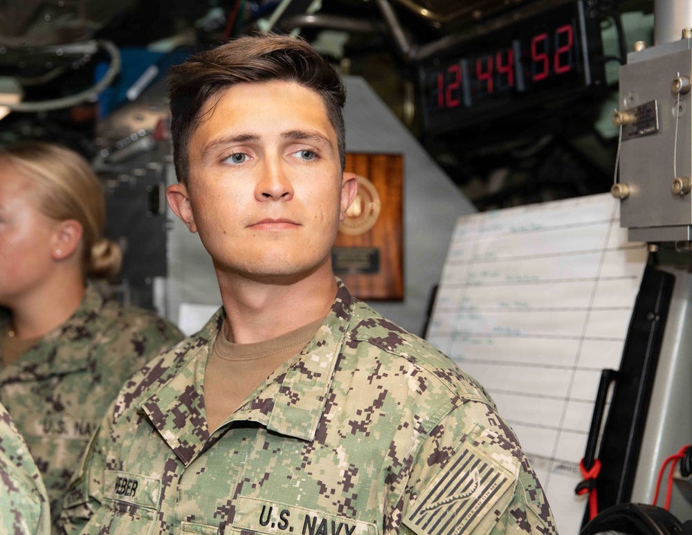 A young U.S. Navy sailor in NWU Type III digital camouflage uniform is standing at attention or on watch inside what appears to be a naval vessel's interior compartment. A digital clock in the background displays 12:44:52, and a whiteboard with handwritten notes is visible nearby. A second sailor in the same uniform is partially visible in the background.