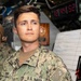 A young U.S. Navy sailor in NWU Type III digital camouflage uniform is standing at attention or on watch inside what appears to be a naval vessel's interior compartment. A digital clock in the background displays 12:44:52, and a whiteboard with handwritten notes is visible nearby. A second sailor in the same uniform is partially visible in the background.