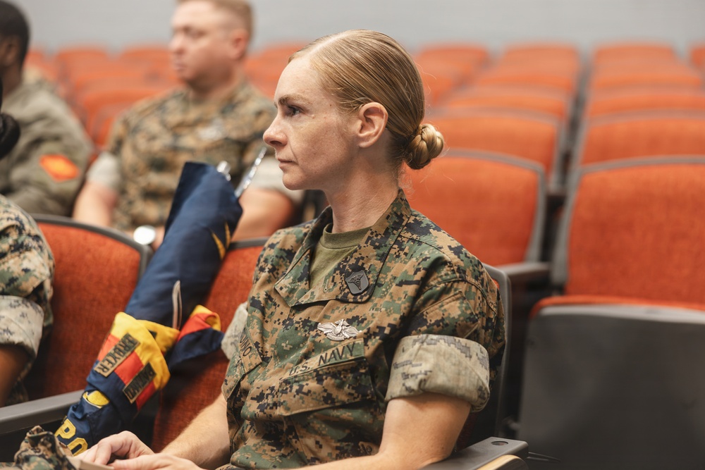 A U.S. Navy service member in Marine Corps MARPAT camouflage uniform is seated in an auditorium-style venue, appearing to attend a briefing or presentation. She wears visible insignia including a U.S. Navy tape, a medical/command badge, and what appears to be a Fleet Marine Force (FMF) device. Other military personnel in similar uniforms are visible in the background.