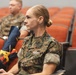 A U.S. Navy service member in Marine Corps MARPAT camouflage uniform is seated in an auditorium-style venue, appearing to attend a briefing or presentation. She wears visible insignia including a U.S. Navy tape, a medical/command badge, and what appears to be a Fleet Marine Force (FMF) device. Other military personnel in similar uniforms are visible in the background.
