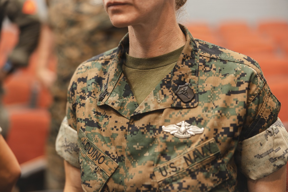 A close-up photograph of a U.S. Navy service member wearing Marine Corps MARPAT digital camouflage utilities. The individual's name tape reads 'AQUINO' and a 'U.S. NAVY' tape is visible on the uniform. Several military insignia and badges are pinned to the uniform, indicating a medical or specialized role.