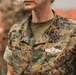 A close-up photograph of a U.S. Navy service member wearing Marine Corps MARPAT digital camouflage utilities. The individual's name tape reads 'AQUINO' and a 'U.S. NAVY' tape is visible on the uniform. Several military insignia and badges are pinned to the uniform, indicating a medical or specialized role.