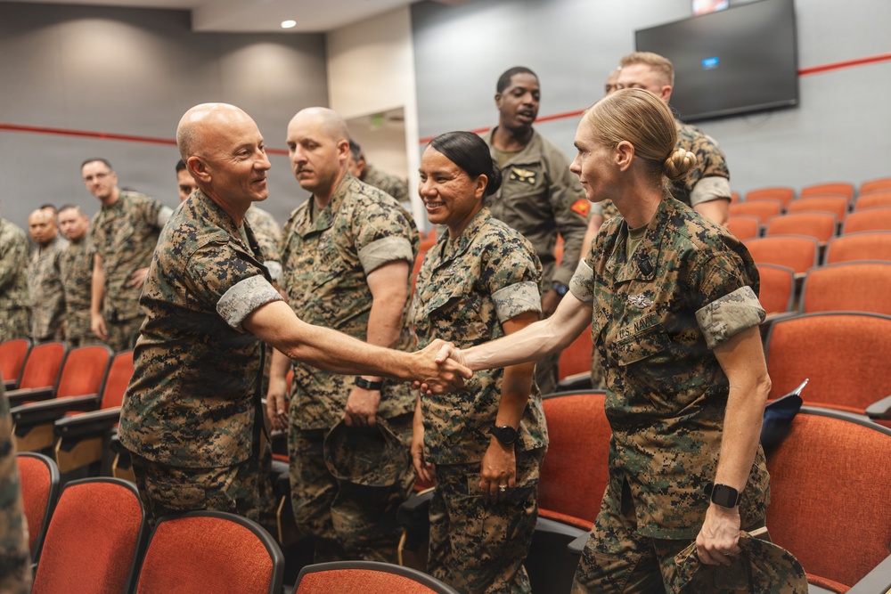 A group of U.S. military personnel in MARPAT camouflage uniforms are gathered in what appears to be a briefing room or auditorium with red/orange seating. Two individuals — a bald male and a female service member with 'U.S. Navy' visible on her uniform — are shaking hands in the foreground. The setting suggests a formal or semi-formal military event such as a meeting, briefing, or recognition ceremony.