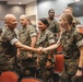A group of U.S. military personnel in MARPAT camouflage uniforms are gathered in what appears to be a briefing room or auditorium with red/orange seating. Two individuals — a bald male and a female service member with 'U.S. Navy' visible on her uniform — are shaking hands in the foreground. The setting suggests a formal or semi-formal military event such as a meeting, briefing, or recognition ceremony.