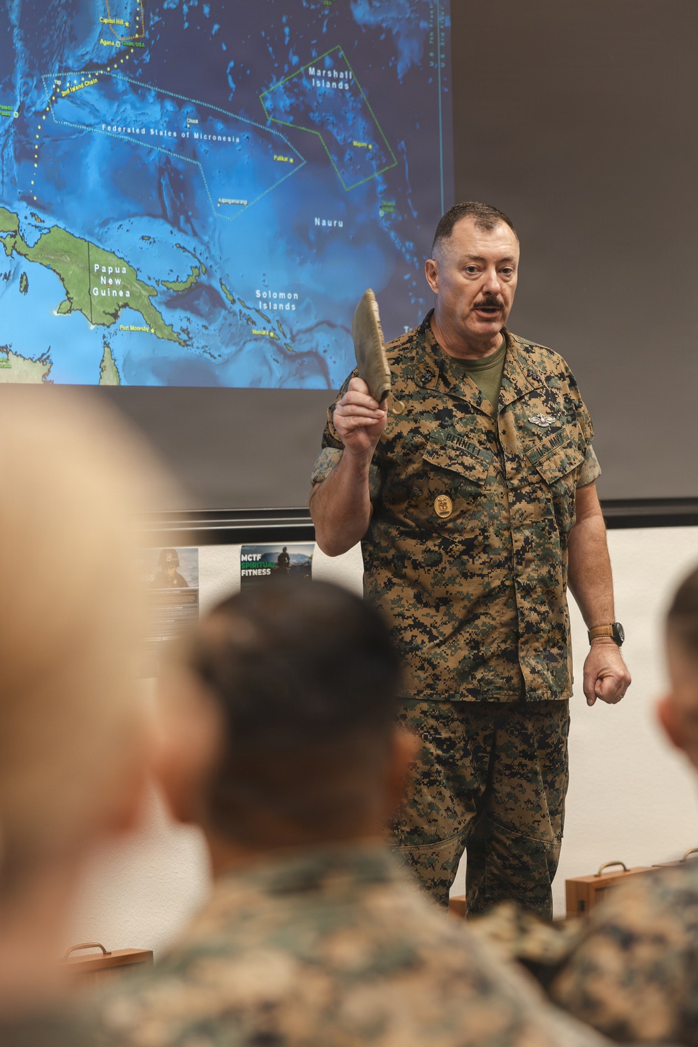 A U.S. Marine Corps officer in MARPAT digital camouflage uniform is delivering a briefing or presentation to an audience. He is holding up what appears to be a tan/khaki boot or shoe as a visual aid, standing in front of a projected map showing the Pacific region including Papua New Guinea, Solomon Islands, Marshall Islands, Nauru, and the Federated States of Micronesia. Several uniformed personnel are visible in the foreground as audience members.