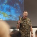 A U.S. Marine Corps officer in MARPAT digital camouflage uniform is delivering a briefing or presentation to an audience. He is holding up what appears to be a tan/khaki boot or shoe as a visual aid, standing in front of a projected map showing the Pacific region including Papua New Guinea, Solomon Islands, Marshall Islands, Nauru, and the Federated States of Micronesia. Several uniformed personnel are visible in the foreground as audience members.