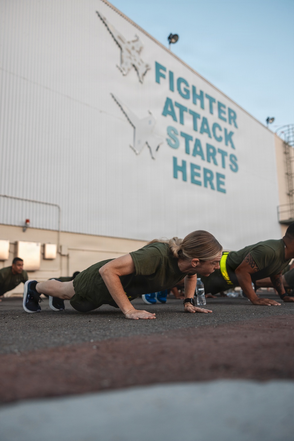 A group of military personnel in olive/dark green PT (physical training) uniforms are performing push-ups on an outdoor tarmac or flight line surface. Behind them is a large white hangar wall painted with the text 'FIGHTER ATTACK STARTS HERE' alongside a mural of a military jet aircraft. The foreground subject is a blonde female service member in sharp focus, with others visible in the background.