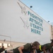 A group of military personnel in olive/dark green PT (physical training) uniforms are performing push-ups on an outdoor tarmac or flight line surface. Behind them is a large white hangar wall painted with the text 'FIGHTER ATTACK STARTS HERE' alongside a mural of a military jet aircraft. The foreground subject is a blonde female service member in sharp focus, with others visible in the background.