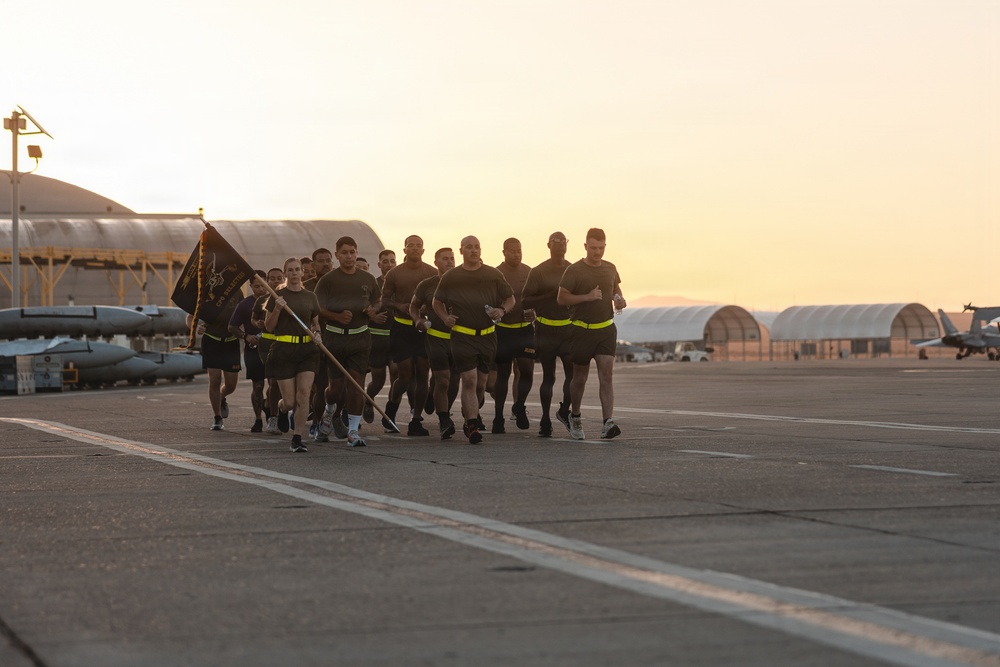 A formation of military personnel in physical training (PT) uniforms are conducting a group run on an airfield tarmac at sunrise or sunset. One individual at the front carries a unit guidon flag. Aircraft hangars and parked military aircraft are visible in the background.