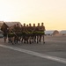 A formation of military personnel in physical training (PT) uniforms are conducting a group run on an airfield tarmac at sunrise or sunset. One individual at the front carries a unit guidon flag. Aircraft hangars and parked military aircraft are visible in the background.