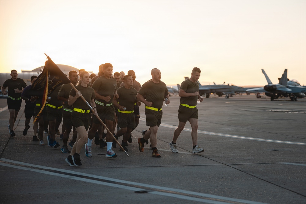 A group of military personnel in uniform are conducting a formation run on an airfield tarmac during early morning or evening hours, as indicated by the golden-hour lighting. One runner carries a unit guidon flag while the group wears matching dark PT (physical training) uniforms with high-visibility yellow reflective belts. Military jet aircraft, appearing to be F/A-18 Hornets or similar fighters, are visible parked on the flight line in the background.