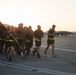 A group of military personnel in uniform are conducting a formation run on an airfield tarmac during early morning or evening hours, as indicated by the golden-hour lighting. One runner carries a unit guidon flag while the group wears matching dark PT (physical training) uniforms with high-visibility yellow reflective belts. Military jet aircraft, appearing to be F/A-18 Hornets or similar fighters, are visible parked on the flight line in the background.