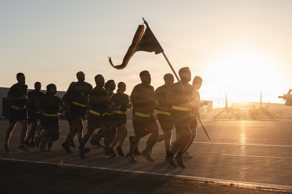 A group of military personnel are conducting a formation run at sunrise or sunset on what appears to be a military installation or airfield. One individual at the front carries a guidon (unit flag) while the group runs in formation wearing reflective safety belts. The dramatic backlighting creates strong silhouettes against a golden sky.