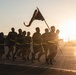 A group of military personnel are conducting a formation run at sunrise or sunset on what appears to be a military installation or airfield. One individual at the front carries a guidon (unit flag) while the group runs in formation wearing reflective safety belts. The dramatic backlighting creates strong silhouettes against a golden sky.