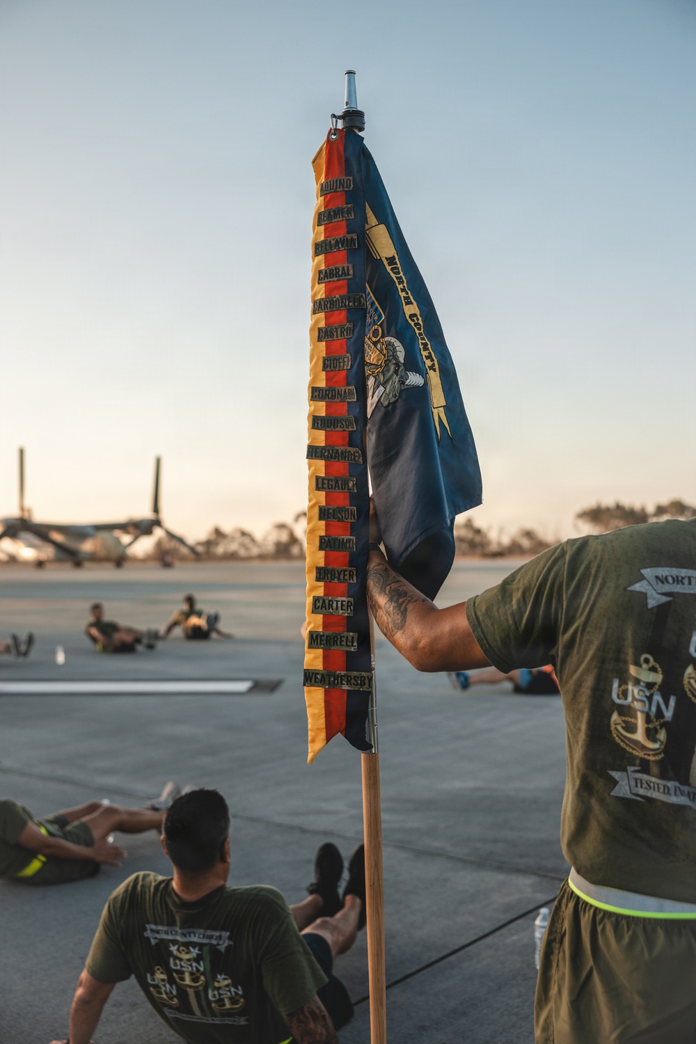 A USN (United States Navy) guidon flag labeled 'North County' is being held upright on a wooden pole during what appears to be a physical training session on a tarmac or airfield. The flag displays multiple name tapes sewn vertically along red and gold streamers, listing individual sailors' surnames. Personnel in olive green USN-branded t-shirts are visible performing exercises in the background.