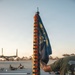 A USN (United States Navy) guidon flag labeled 'North County' is being held upright on a wooden pole during what appears to be a physical training session on a tarmac or airfield. The flag displays multiple name tapes sewn vertically along red and gold streamers, listing individual sailors' surnames. Personnel in olive green USN-branded t-shirts are visible performing exercises in the background.