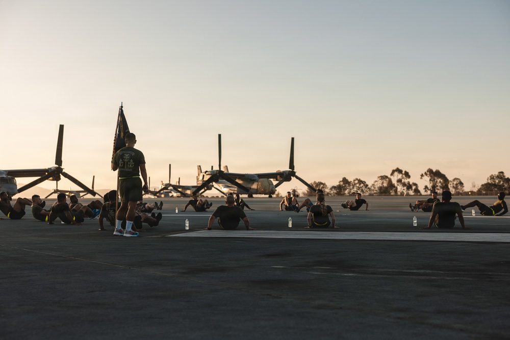A group of military personnel are conducting physical training exercises on an airfield tarmac during early morning or evening golden hour lighting. One individual stands holding a unit guidon/flag while others perform sit-ups or crunches on the ground. Several V-22 Osprey tiltrotor aircraft are parked in the background.