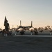 A group of military personnel are conducting physical training exercises on an airfield tarmac during early morning or evening golden hour lighting. One individual stands holding a unit guidon/flag while others perform sit-ups or crunches on the ground. Several V-22 Osprey tiltrotor aircraft are parked in the background.