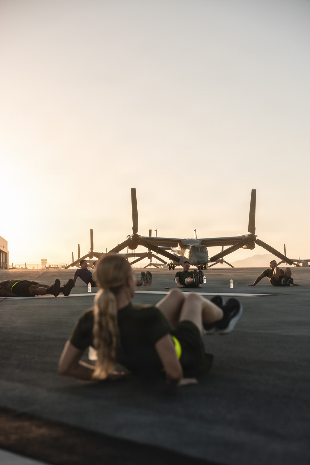 A group of military personnel are performing physical training exercises on a flight line tarmac at either sunrise or sunset. They are doing sit-ups and other floor exercises in front of a parked V-22 Osprey tiltrotor aircraft. The warm golden light and mountain silhouettes in the background suggest a desert military installation.