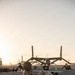 A group of military personnel are performing physical training exercises on a flight line tarmac at either sunrise or sunset. They are doing sit-ups and other floor exercises in front of a parked V-22 Osprey tiltrotor aircraft. The warm golden light and mountain silhouettes in the background suggest a desert military installation.