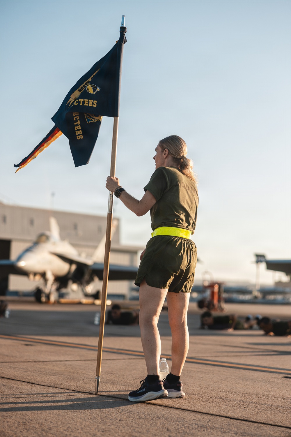 A person in military physical training attire holds a dark blue guidon flag on an airfield tarmac. The flag displays text including 'CTEES' and gold insignia, consistent with a military unit guidon. In the background, a military jet aircraft and hangar structures are visible, along with other personnel performing exercises on the ground.