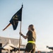A person in military physical training attire holds a dark blue guidon flag on an airfield tarmac. The flag displays text including 'CTEES' and gold insignia, consistent with a military unit guidon. In the background, a military jet aircraft and hangar structures are visible, along with other personnel performing exercises on the ground.