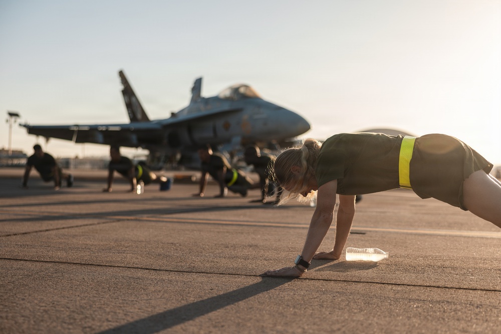 Multiple individuals are performing push-ups on a military airfield tarmac in front of a parked fighter jet, likely an F/A-18 Hornet or Super Hornet. The foreground subject has long hair pulled back and wears olive/dark green athletic attire with a yellow-green reflective waistband. The scene appears to be an early morning or late afternoon physical training session based on the low-angle, warm sunlight.