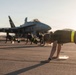 Multiple individuals are performing push-ups on a military airfield tarmac in front of a parked fighter jet, likely an F/A-18 Hornet or Super Hornet. The foreground subject has long hair pulled back and wears olive/dark green athletic attire with a yellow-green reflective waistband. The scene appears to be an early morning or late afternoon physical training session based on the low-angle, warm sunlight.