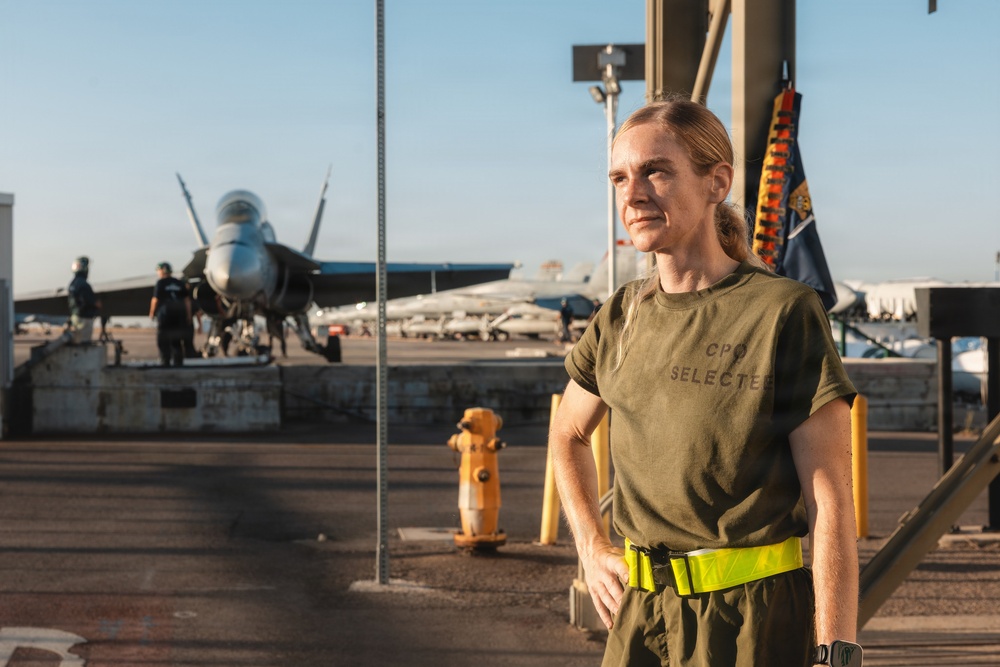 A person wearing an olive green 'CPO SELECTEE' t-shirt and a high-visibility yellow belt stands on what appears to be a naval aircraft carrier flight deck. A military jet aircraft, likely an F/A-18 Hornet or Super Hornet, is visible in the background along with other personnel and deck equipment. The image appears to be a portrait-style photograph taken during golden hour lighting.