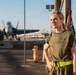 A person wearing an olive green 'CPO SELECTEE' t-shirt and a high-visibility yellow belt stands on what appears to be a naval aircraft carrier flight deck. A military jet aircraft, likely an F/A-18 Hornet or Super Hornet, is visible in the background along with other personnel and deck equipment. The image appears to be a portrait-style photograph taken during golden hour lighting.