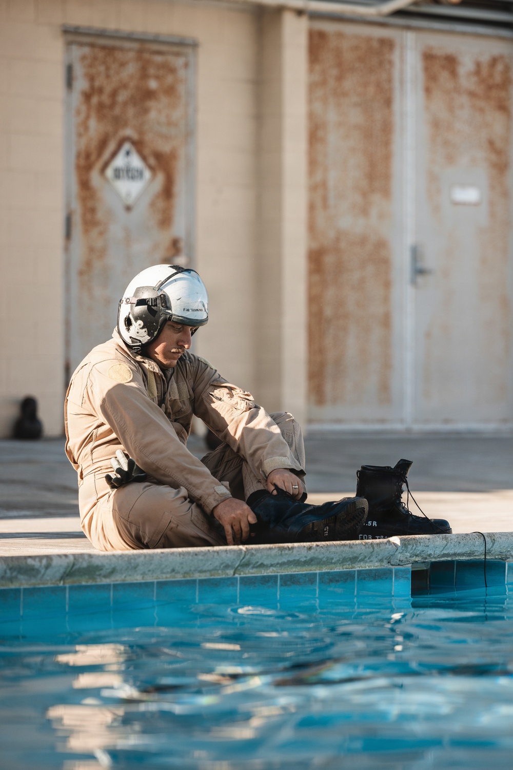 A military aviator in a tan flight suit and white flight helmet is seated at the edge of a pool, removing or adjusting black combat boots. The setting appears to be a military installation, with weathered/rusted metal doors visible in the background. The scene suggests a water survival or egress training exercise.