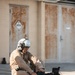 A military aviator in a tan flight suit and white flight helmet is seated at the edge of a pool, removing or adjusting black combat boots. The setting appears to be a military installation, with weathered/rusted metal doors visible in the background. The scene suggests a water survival or egress training exercise.