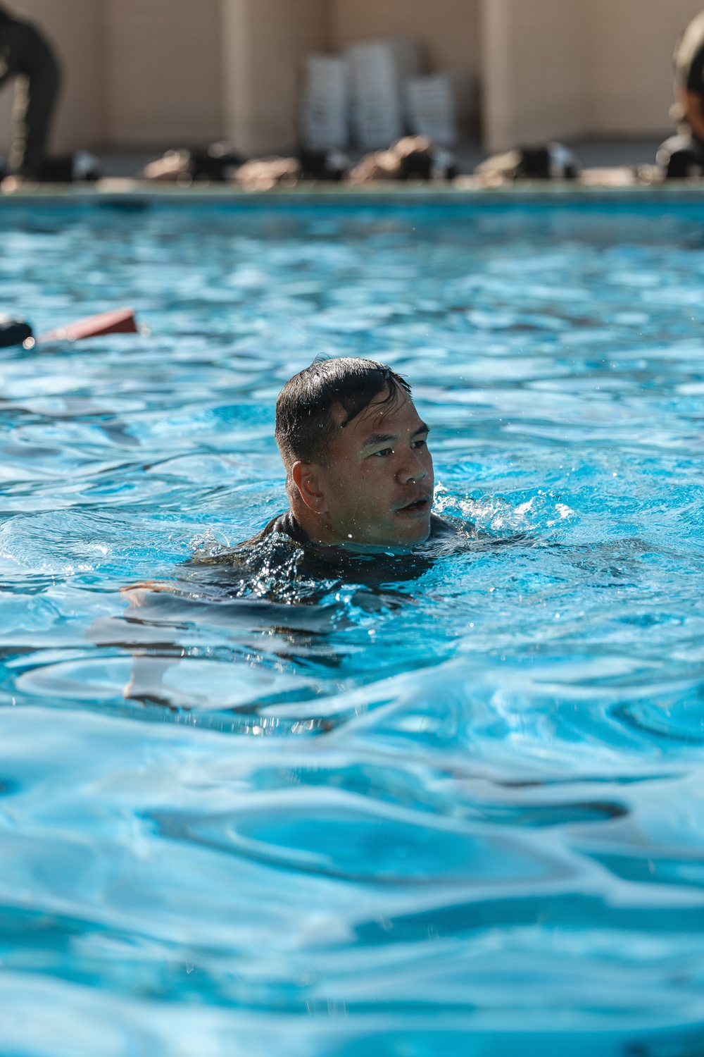 A man is swimming in a blue outdoor pool, with his head and upper shoulders visible above the water surface. He appears to be wearing a dark-colored garment, possibly a wetsuit or uniform, suggesting this may be a military or training exercise. Several observers or participants are visible seated along the pool edge in the background.