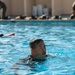 A man is swimming in a blue outdoor pool, with his head and upper shoulders visible above the water surface. He appears to be wearing a dark-colored garment, possibly a wetsuit or uniform, suggesting this may be a military or training exercise. Several observers or participants are visible seated along the pool edge in the background.