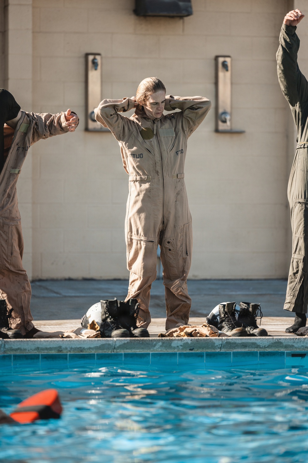 The image shows military personnel in flight suits standing at the edge of an outdoor pool, apparently participating in water survival training. The central figure, wearing a tan/khaki flight suit with 'FTU' markings, has wet hair and appears to have recently exited the water. Multiple pairs of military boots and helmets are visible on the pool deck.