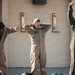The image shows military personnel in flight suits standing at the edge of an outdoor pool, apparently participating in water survival training. The central figure, wearing a tan/khaki flight suit with 'FTU' markings, has wet hair and appears to have recently exited the water. Multiple pairs of military boots and helmets are visible on the pool deck.