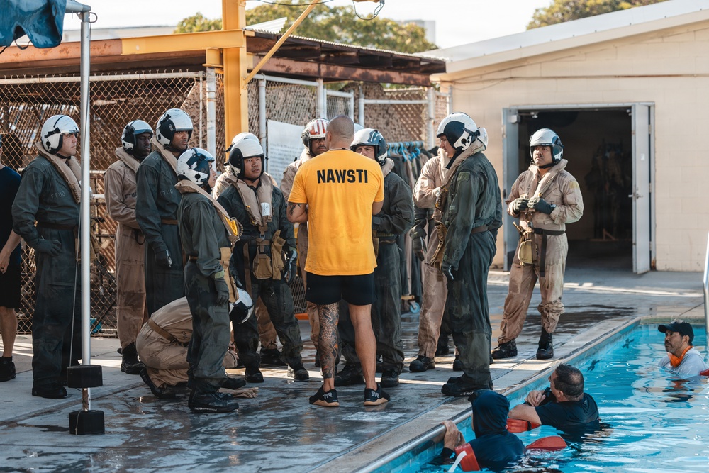 A NAWSTI (Naval Aviation Water Survival Training Instructor) in a yellow shirt addresses a group of military personnel wearing flight suits and white helmets near a training pool. The scene depicts a water survival training exercise at what appears to be a military aquatic training facility. Two individuals are already in the pool, suggesting active training is underway.