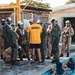 A NAWSTI (Naval Aviation Water Survival Training Instructor) in a yellow shirt addresses a group of military personnel wearing flight suits and white helmets near a training pool. The scene depicts a water survival training exercise at what appears to be a military aquatic training facility. Two individuals are already in the pool, suggesting active training is underway.