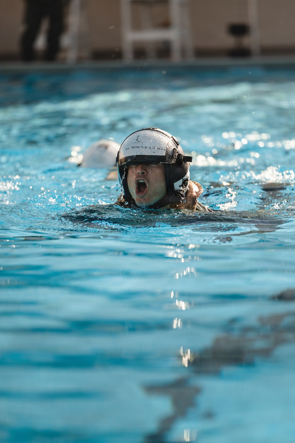 A person wearing a military or aviation-style helmet is swimming in an indoor pool with their mouth open, appearing to be in a training exercise. The swimmer is partially submerged with only their head and upper body visible above the waterline. The setting appears to be a controlled aquatic training environment, likely a survival or water egress training exercise.