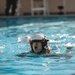 A person wearing a military or aviation-style helmet is swimming in an indoor pool with their mouth open, appearing to be in a training exercise. The swimmer is partially submerged with only their head and upper body visible above the waterline. The setting appears to be a controlled aquatic training environment, likely a survival or water egress training exercise.