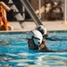A person wearing a white flight helmet and what appears to be a flight suit is swimming in a pool, likely during a water survival training exercise. Water is dripping from the helmet and the individual's mouth is open, suggesting exertion. A second person in similar gear is visible in the background, and safety equipment including an orange life vest is visible poolside.