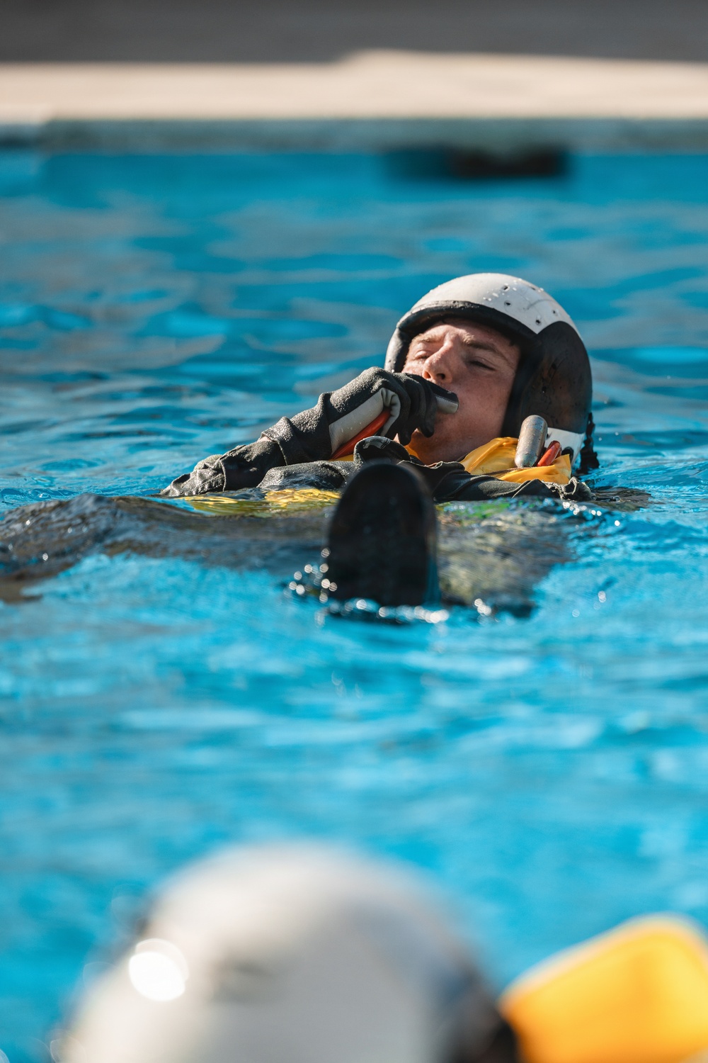 A person wearing a flight helmet, black gloves, and a yellow inflated life preserver vest is floating in a pool of blue water. They appear to be blowing into an inflation tube on their survival vest, suggesting military or aviation water survival training. A second partially visible person in similar gear is seen in the foreground.