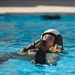 A person wearing a flight helmet, black gloves, and a yellow inflated life preserver vest is floating in a pool of blue water. They appear to be blowing into an inflation tube on their survival vest, suggesting military or aviation water survival training. A second partially visible person in similar gear is seen in the foreground.