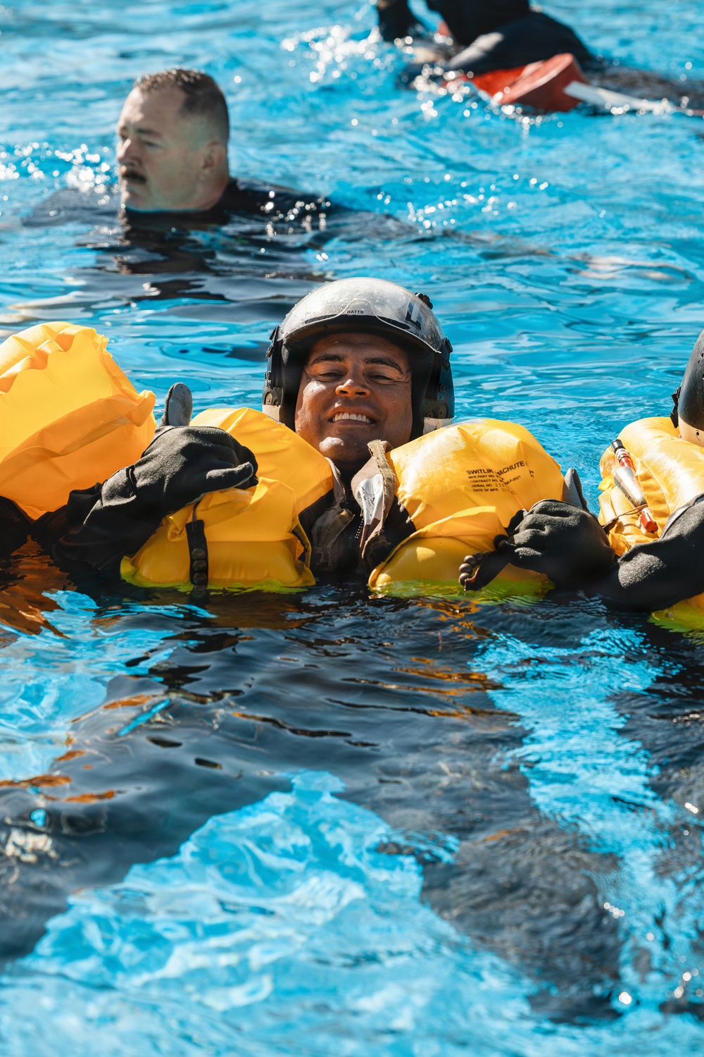 Multiple individuals are in a pool of water during what appears to be a military or aviation water survival training exercise. The central figure is wearing a flight helmet and an inflated yellow life preserver unit (LPU), smiling broadly while treading water. Additional participants are visible in the background, one wearing a black wetsuit and another with an orange flotation device.