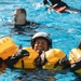 Multiple individuals are in a pool of water during what appears to be a military or aviation water survival training exercise. The central figure is wearing a flight helmet and an inflated yellow life preserver unit (LPU), smiling broadly while treading water. Additional participants are visible in the background, one wearing a black wetsuit and another with an orange flotation device.