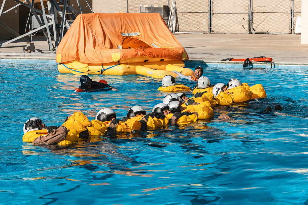 A water survival training exercise is taking place in an outdoor pool facility. Multiple trainees wearing white helmets and yellow inflatable life vests are linked together in a chain formation in the water, practicing survival techniques. An orange inflatable life raft is deployed in the background of the pool.