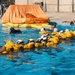 A water survival training exercise is taking place in an outdoor pool facility. Multiple trainees wearing white helmets and yellow inflatable life vests are linked together in a chain formation in the water, practicing survival techniques. An orange inflatable life raft is deployed in the background of the pool.