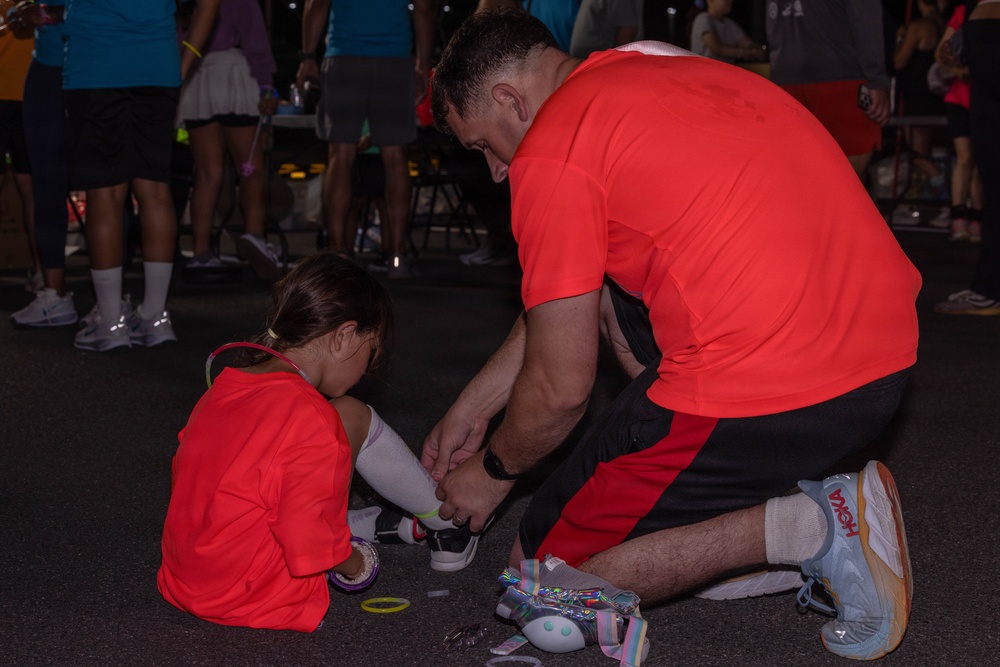 An adult male in a red shirt kneels on the ground to help a young girl, also wearing a red shirt, with her footwear or roller skates. The scene appears to take place at a nighttime outdoor event with a crowd of people visible in the background. Both individuals are wearing matching red shirts, suggesting they may be part of the same team or event group.