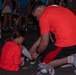 An adult male in a red shirt kneels on the ground to help a young girl, also wearing a red shirt, with her footwear or roller skates. The scene appears to take place at a nighttime outdoor event with a crowd of people visible in the background. Both individuals are wearing matching red shirts, suggesting they may be part of the same team or event group.