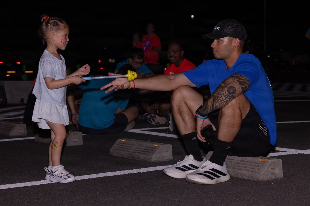 A young girl with face paint and colorful stickers on her leg is handing or receiving a glow stick from a tattooed adult male crouching on a parking lot curb stop at night. The scene appears to be an outdoor nighttime event, likely a fun run or glow-themed community gathering. Several other participants are visible in the background, some wearing glow accessories.